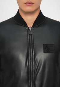 Personne portant une veste en cuir noire avec une fermeture éclair à l'avant et un patch logo Armani Exchange sur la poitrine, col visible, lèvres et menton visibles.