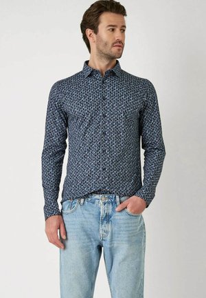 Marineblauw shirt met lange mouwen en een geometrisch patroon in lichtblauw en wit. Met knoopsluiting en slim fit. Gepaard met lichtblauwe jeans.