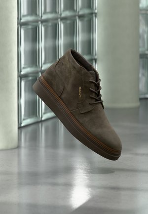 Brauner Wildleder-Schnürstiefelette mit dicker Sohle, schwebend über einem polierten Boden nahe Glasblockwänden gezeigt.