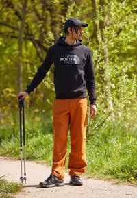 Homme en sweat à capuche noir et pantalons orange tenant des bâtons de randonnée se tient sur un chemin de terre dans une forêt verte, regardant sur le côté.