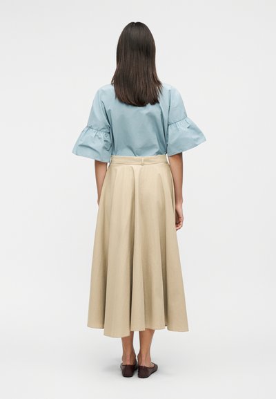 Γυναίκα με μακριά σκούρα μαλλιά στέκεται γυρισμένη πίσω, φορώντας ανοιχτό μπλε μπλούζα με φουσκωτά μανίκια, μπεζ midi φούστα και σκούρα επίπεδα παπούτσια.