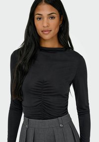 Kvinne med langt svart hår iført en svart rynket langermet topp og høy inntrekket grå plissert bukser foran en nøytral bakgrunn.