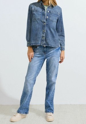Camisa de mezclilla en azul medio con mangas largas, botones en la parte delantera y dos bolsillos en el pecho; combinada con jeans rectos de color azul claro y zapatillas beiges.