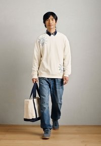 Άνδρας περπατάει σε εσωτερικό χώρο φορώντας ένα άσπρο φούτερ με σχέδια ήλιου, μπλε τζιν, γκρι παπούτσια και κρατώντας μια μπεζ tote bag με σκούρες λαβές.