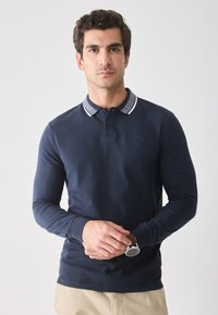 Marineblå langærmet polo med mønstret krave, knaplukning og broderet logo. Modellen bærer et ur og beige bukser.