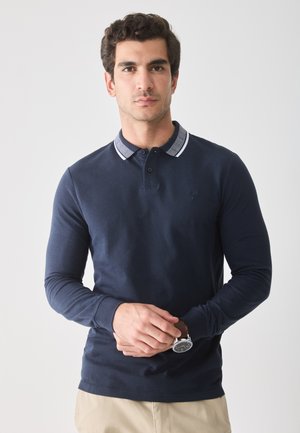 Mornarice dugih rukava polo majica s oštrim okovratnikom, gumbima i vezom logotipa. Model nosi sat i bež hlače.