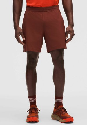 Mand iført rustfarvede atletikshorts, matchende sokker med hvide striber og orange løbesko står mod en ensfarvet baggrund.