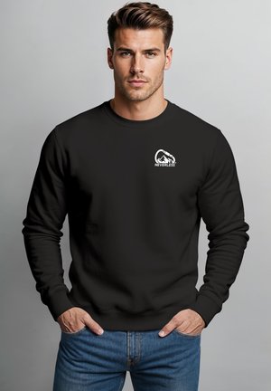 Mann trägt schwarzen Sweatshirt mit Bergkarabiner-Logo und "NEVERLESS"-Text, Hände in den Taschen, steht vor schlichtem grauem Hintergrund.