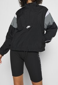 Svart nylonpullovjacka med grå och vita panelaccentuerade detaljer, halvdragkedja, elastiska ärmar och dragsko i nederkant. Nike-logotyp på framsidan.