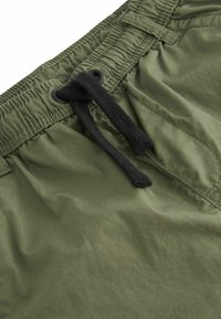 Pantaloni da bagno verde oliva con vita elastica arricciata, cordino nero e una texture liscia e leggera. Presentano dettagli di cucitura sottili.
