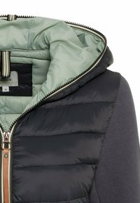 Veste matelassée noire avec doublure intérieure vert clair, fermeture éclair avec tirette beige et manches en tissu gris foncé.