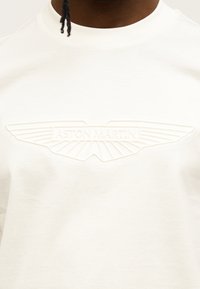 Λευκό βαμβακερό μπλουζάκι με ανάγλυφο λογότυπο Aston Martin και σχεδιασμό φτερού στο μπροστινό μέρος. Ομαλή υφή και κλασικό στρογγυλό λαιμό.