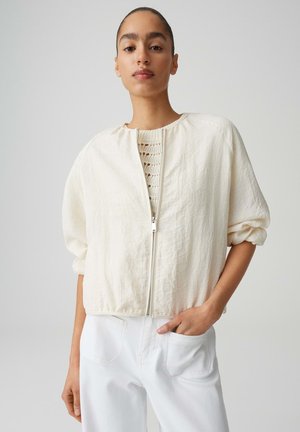 Persona giovane con capelli pettinati all'indietro che indossa una giacca crema testurizzata con zip sopra una maglia lavorata a maglia e pantaloni bianchi, in piedi davanti a uno sfondo semplice.