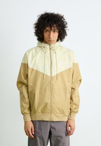 Beige and cream windbreaker with a hood, featuring a chevron pattern, zip closure, and drawstring. Includes a small white logo on the chest.