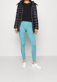 Vadderad svart jacka, svart topp och turkosa leggings med ormmönster. Vita sneakers kompletterar outfiten och framhäver de livfulla färgerna och mönstren.
