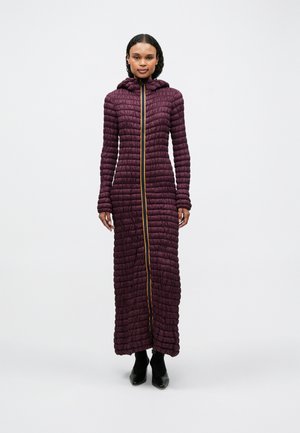Dlhý, fialový, prešívaný kabát s kapucňou, ktorý má zlatý plný zips vpredu. Textúra je nadýchaná s vertikálnym prešívaním. Čierna obuv.