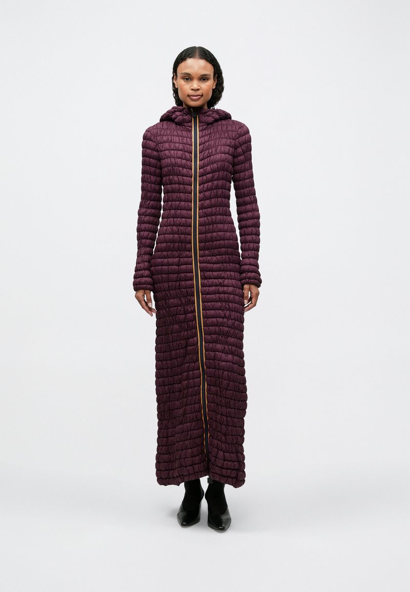 Μακρύ, μωβ, επενδεδυμένο παλτό με κουκούλα, που διαθέτει πλήρη εμπρόσθιο φερμουάρ σε χρυσό. Η υφή είναι φουσκωτή με κατακόρυφες ραφές. Μαύρα υποδήματα.