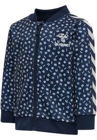 Navyblauwe ritsjack met witte geometrische patronen, geribde manchetten en een sportieve streepdetail aan de zijkanten. Zacht materiaal, ontspannen pasvorm.
