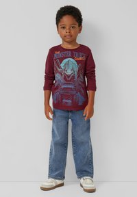 Bordeauxrotes Langarm-T-Shirt mit einem Grafikdesign eines Monstertrucks und eines Dinosauriers. Getragen mit blauen Jeans und weißen Sneakers.
