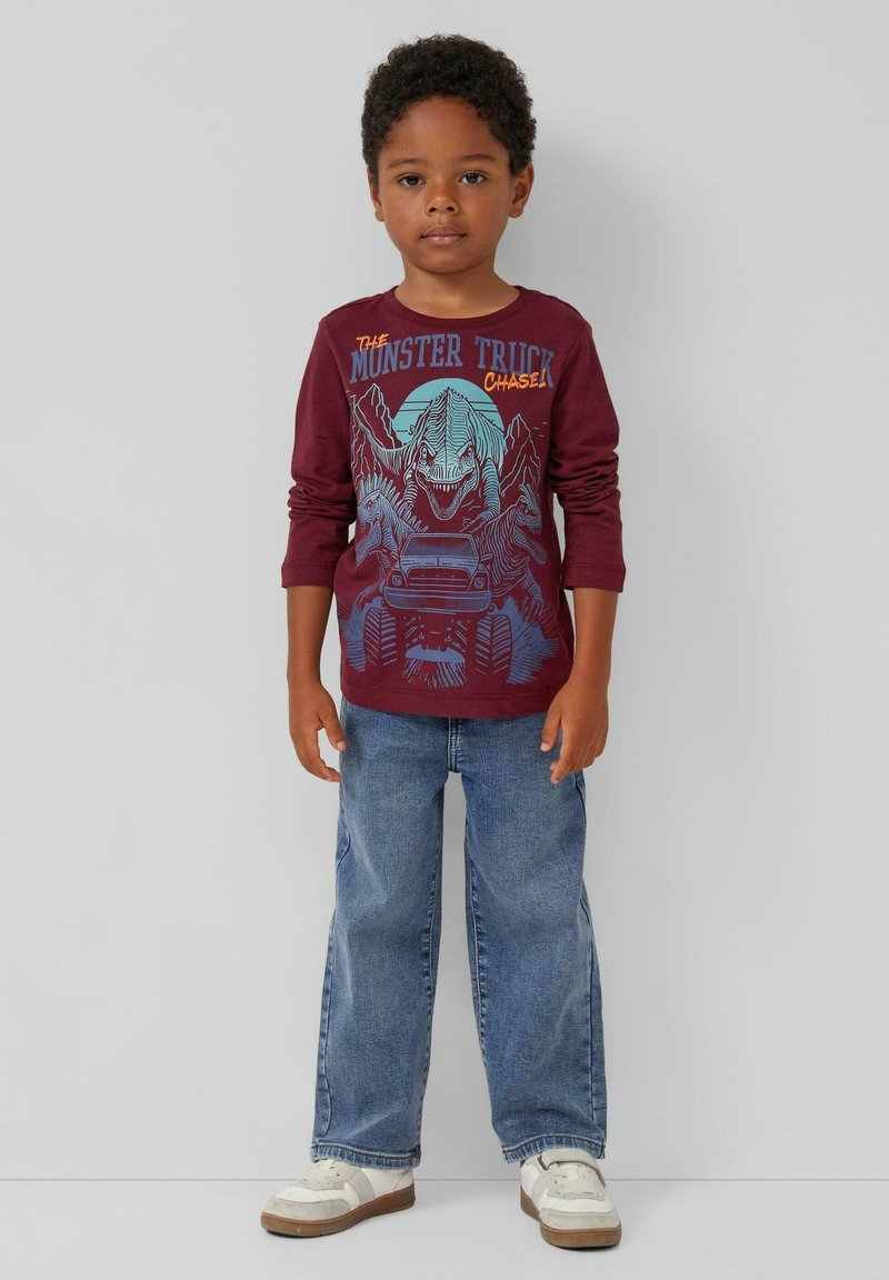 Bordeauxrotes Langarm-T-Shirt mit einem Grafikdesign eines Monstertrucks und eines Dinosauriers. Getragen mit blauen Jeans und weißen Sneakers.