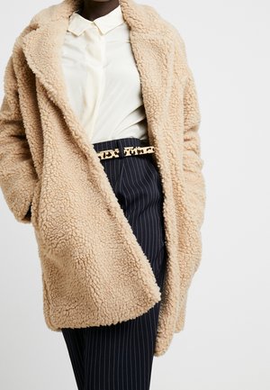 Personne portant un manteau teddy beige, une chemise blanche à col et un pantalon à rayures bleu marine avec une ceinture motif léopard sur un fond uni.