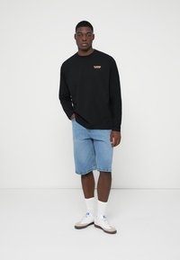 Cămașă neagră cu mâneci lungi și logo portocaliu, asociată cu pantaloni scurți din denim bleu deschis. Șosete albe și adidași completează ținuta.
