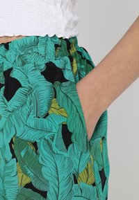Pantalons turquoise avec un motif de feuilles tropicales en vert et noir. Taille élastique avec poches latérales, fabriqués en tissu léger.