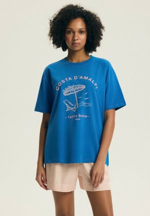 Kobieta o kręconych włosach, ubrana w niebieską koszulkę "Costa D'Amalfi" z nadrukiem plaży oraz jasne, paskowane szorty na jednolitym tle.