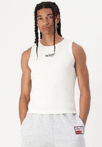 Valge soonikust varrukateta top, millel on ümar kaelus ja rinna kohal must tikandiga logo. Modell kannab halli dressipükse plaastriga.