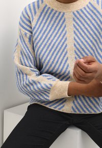 Pull en tricot avec une base beige et des rayures diagonales bleues, col et poignets à côtes, et coupe décontractée. Main d'une personne visible.