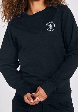 Personne portant une chemise à manches longues noire de U.S. Polo Assn. avec le logo sur le côté gauche de la poitrine, mains jointes devant, cheveux bouclés partiellement visibles.