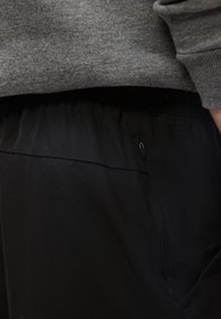 Άτομο που φοράει μαύρο παντελόνι με τσέπη με φερμουάρ και γκρι μακρυμάνικη μπλούζα, δείχνοντας την περιοχή της μέσης και του χεριού.