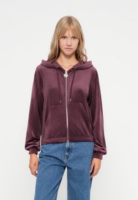 Bordeaux kleurige ritssluiting hoodie in zachte velours met een gevoerde capuchon, voorzakken en aantrekkoord details. Gekoeld met lichtblauwe spijkerbroek.