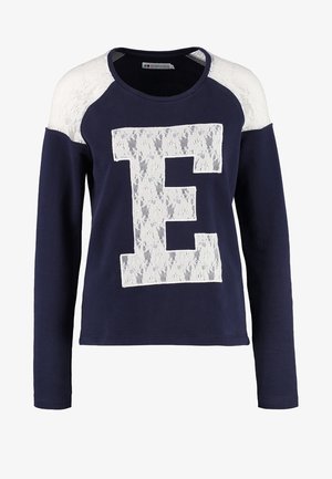 Marineblå sweater med hvide blonder, stort "E" grafisk design og lange ærmer, parret med lyseblå jeans og beige ankelstøvler.