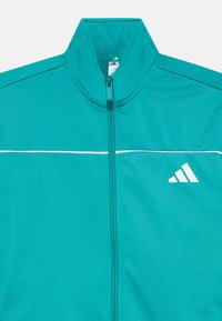 Veste de survêtement teal avec un col montant, fermeture zippée, rayure horizontale blanche et logo blanc sur le côté gauche. Texture de tissu lisse.