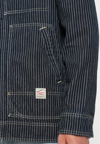 Blue denim jacket featuring white vertical stripes, front pocket, and a logo patch. Textured fabric with visible stitching accents.