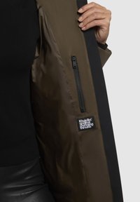 Hand holding open black and olive jacket showing inner zip pocket and fabric label with white text.