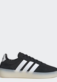 Sneaker nero basso con tre strisce bianche sul lato, lacci neri, linguetta argentata sul tallone e suola traslucida.