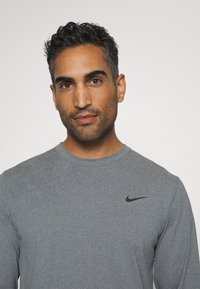 Hosszú ujjú szürke sportpóló kerek nyakkivágással és fekete Nike logóval a bal mellen, sima, texturált anyagból készült.
