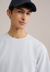 Svetlo siva majica s dugim rukavima sa okruglim izrezom i ležernim krojem, uparena s tamnoplavom kapom. Glatka tekstura i jednostavan dizajn.