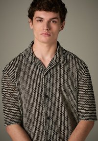 Camicia a maniche corte con bottoni in un motivo a zigzag bianco e nero, con colletto e bottoni neri, realizzata in tessuto intrecciato e strutturato.