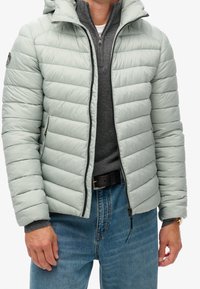 Chaqueta acolchada verde claro con capucha, con diseño acolchado, cremallera frontal y bolsillos laterales, llevada sobre un suéter gris.