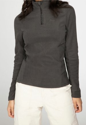Femme portant un pull polaire gris foncé à manches longues avec une courte fermeture éclair devant et un pantalon beige clair sur un fond uni.