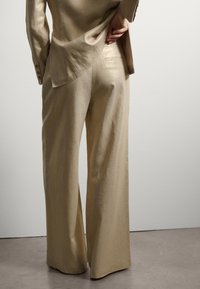 Completo in lino beige con pantaloni a gamba larga. La giacca presenta un orlo curvo e un leggero luccichio, accentuato da una tasca sul retro dei pantaloni.