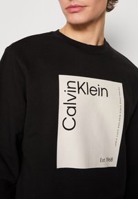 Felpa nera con una grande grafica rettangolare bianca che riporta il testo "Calvin Klein" e "Est. 1968." Design classico a girocollo, materiale in cotone.