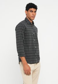 Man die een zwart-wit geblokt overhemd met lange mouwen draagt, met beige broek, staande tegen een effen witte achtergrond.