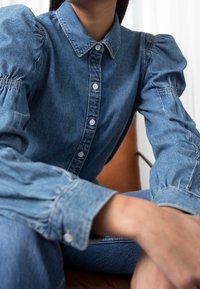 Denim button-up shirt with puff sleeves, light blue color, detailed stitching, and white buttons. Worn with matching jeans.