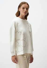 Crèmekleurige sweatshirt met een oversized pasvorm, voorzien van een beige bloemenomtrekontwerp aan de voorkant. Zacht, glad materiaal met ribgebreide manchetten.