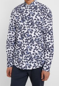 Camicia bianca con un motivo floreale blu, dotata di maniche lunghe e colletto a punta. Realizzata in tessuto leggero.