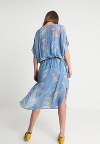 Mujer vestida con un fluido vestido azul con patrones de hojas multicolores, de espaldas a la cámara, combinando con zapatos de tacón amarillos.